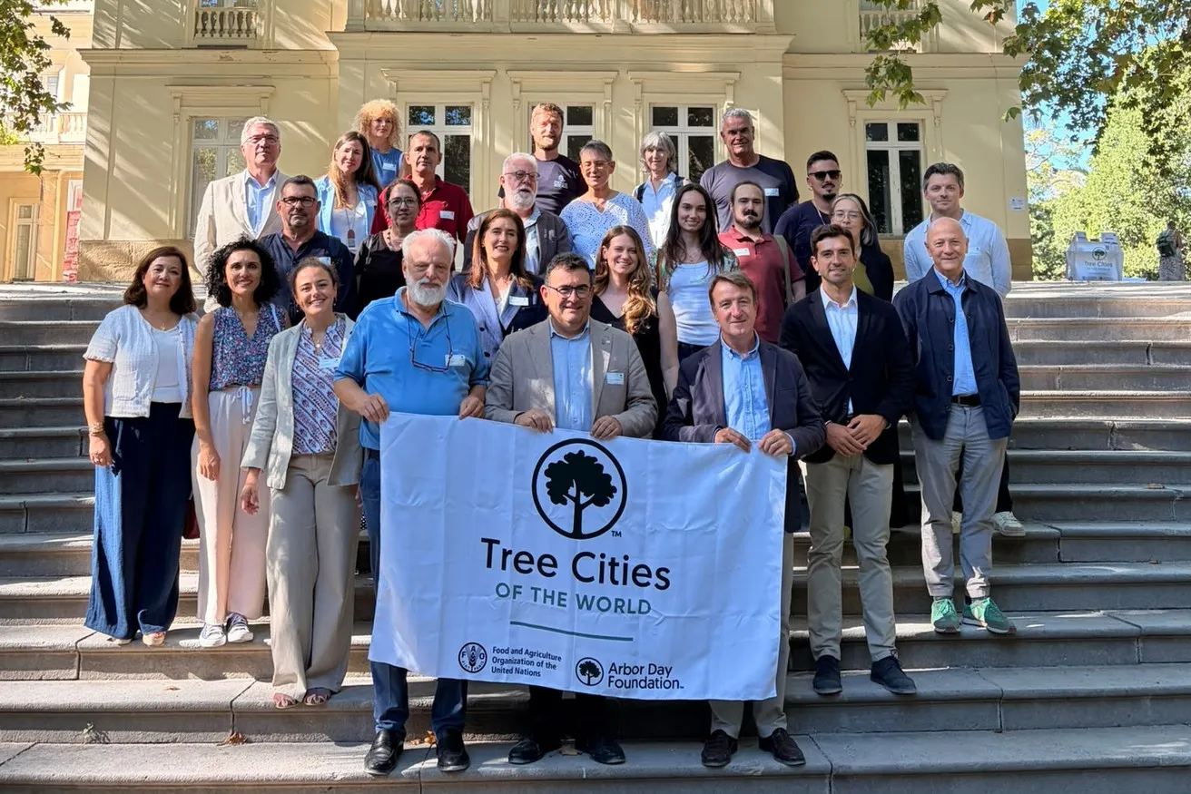 About 25 attendees of the Tree Cities of the World Europe gathering in Málaga pose on stone steps in front of a cream neoclassical building. People in the front row hold a white Tree Cities of the World banner with the FAO and Arbor Day Foundation logos.